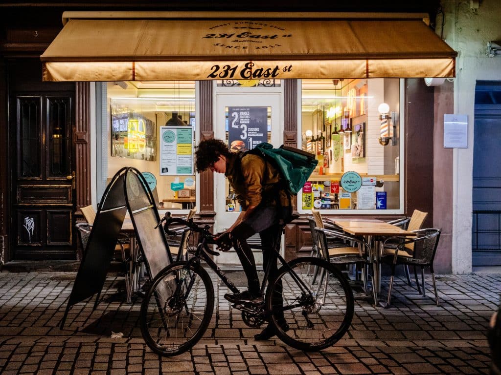 A gig-worker on a bike with an empty bag searches for a job outside of a restaurant.