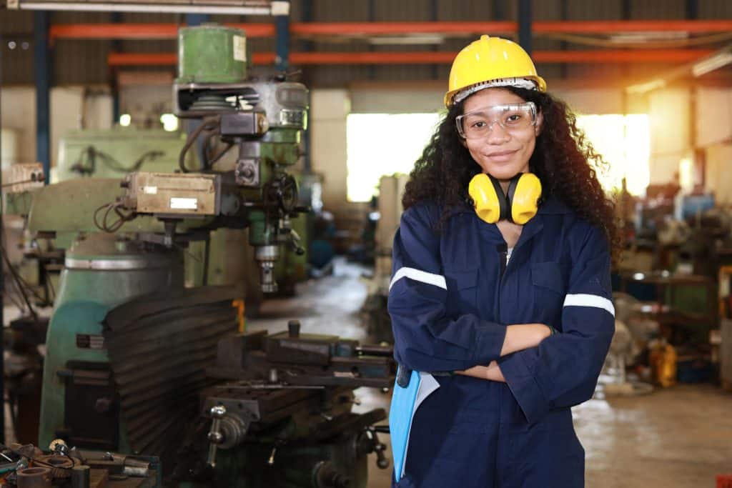 Teen worker with long, dark, curly hair wearing a hardhat, gloves, protective goggles, and protective ear covers folds her arms and smiles in a workshop full of machinery.