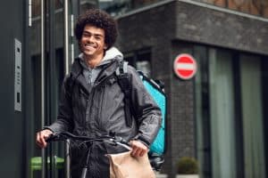 smiling-gig-worker-with-bag-in-hand-and-delivery-box-on-back-atop-bike